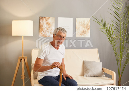 Serious old disabled man holding hands on wooden stick, resting on cozy couch in living room elderly senior grandfather wearing casual clothing looking at camera. Serious old disabled man holding hands on wooden stick, resting on cozy couch in living room elderly senior grandfather wearing casual clothing looking at camera. 107617454