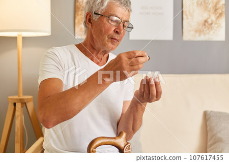 Sick grey-haired mature man wearing casual white T-shirt sitting on sofa at home interior suffering from hypertension, putting drops into glass needs to take medicine. 107617455