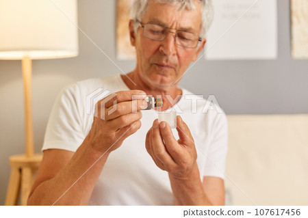 Unhealthy grey-haired senior man wearing casual white T-shirt sitting on couch in living room putting drops into glass time to take medicine feeling unwell needs pills.. Unhealthy grey-haired senior man wearing casual white T-shirt sitting on couch in living room putting drops into glass time to take medicine feeling unwell needs pills.. 107617456