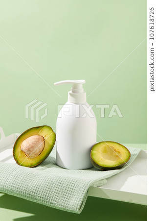 Minimalist concept for advertising cosmetic based on avocado extract. A white pump bottle placed on cotton towel with halves of avocado on pastel background. Empty label for design. Front view Minimalist concept for advertising cosmetic based on avocado extract. A white pump bottle placed on cotton towel with halves of avocado on pastel background. Empty label for design. Front view 107618726