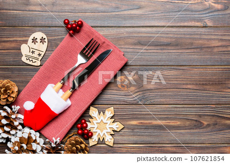 Top view of fork and knife on napkin on wooden background. Different christmas decorations and toys. New Year dinner concept with empty space for your design Top view of fork and knife on napkin on wooden background. Different christmas decorations and toys. New Year dinner concept with empty space for your design 107621854