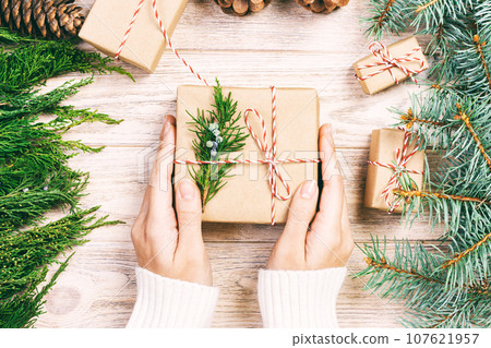 Hand crafted gift on wooden background with Christmas decor. Woman wrapping christmas present , girl prepares xmas gifts with fir tree and pine cone. Top view, copy space. Toned Hand crafted gift on wooden background with Christmas decor. Woman wrapping christmas present , girl prepares xmas gifts with fir tree and pine cone. Top view, copy space. Toned 107621957