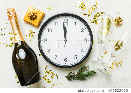 New year celebration concept with a bottle of champagne and two glasses toasting. Christmas gift box, alarm clock and fir tree branch on colored table. Top view Copy space 107622010
