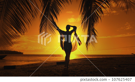Bright color sunset over tropical island beach landscape. Woman silhouette in long shirt playing with palm leaf against orange sky. Outdoor lifestyle travel on summer holiday vacation. Slow motion 4K Bright color sunset over tropical island beach landscape. Woman silhouette in long shirt playing with palm leaf against orange sky. Outdoor lifestyle travel on summer holiday vacation. Slow motion 4K 107625407