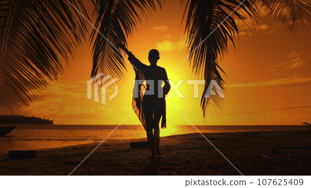 Bright color sunset over tropical island beach landscape. Woman silhouette in long shirt playing with palm leaf against orange sky. Outdoor lifestyle travel on summer holiday vacation. Slow motion 4K 107625409