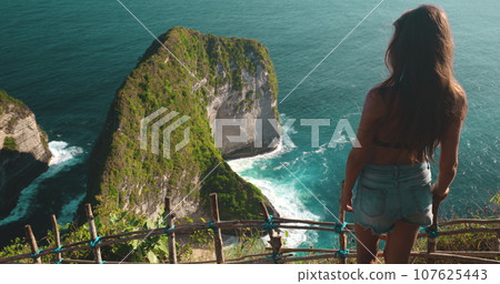 Tourist place to visit - Kelingking Secret Point Beach Indonesia, Nusa Penida Island. Caucasian beautiful woman relax and enjoy aerial ocean mountain wild nature landscape. Slow motion zoom back view Tourist place to visit - Kelingking Secret Point Beach Indonesia, Nusa Penida Island. Caucasian beautiful woman relax and enjoy aerial ocean mountain wild nature landscape. Slow motion zoom back view 107625443