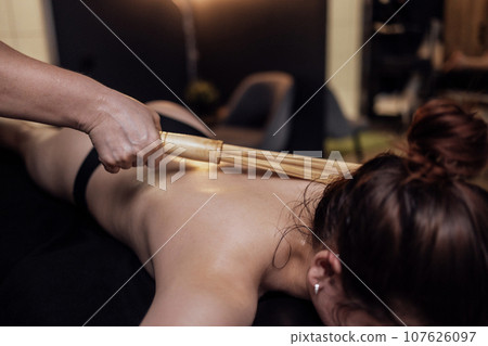 Masseuse makes back massage with a bamboo sticks in the spa salon. Cervical collar area massage with bamboo broom in beauty centre or clinic. 107626097