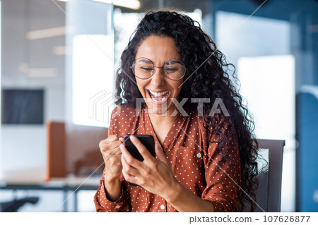 Young beautiful hispanic woman with curly hair inside office celebrating victory success, businesswoman received win notification on phone, using smartphone app at workplace. Young beautiful hispanic woman with curly hair inside office celebrating victory success, businesswoman received win notification on phone, using smartphone app at workplace. 107626877