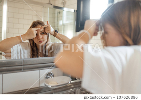 Cute teenage girl squeezes out a pimple on her cheek in the bathroom 107626886
