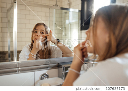 Cute teenage girl squeezes out a pimple on her cheek in the bathroom 107627173