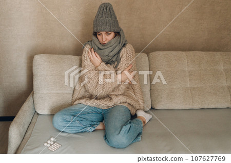 Young woman wearing autumn winter scarf and wool hat has flu ill sick disease cold at home indoor on sofa Young woman wearing autumn winter scarf and wool hat has flu ill sick disease cold at home indoor on sofa 107627769