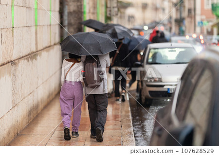 Two girls walking down the street under an umbrella in the rain 107628529