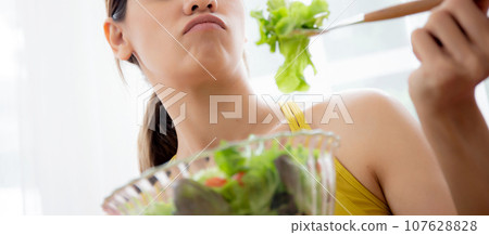 Young caucasian woman sitting on sofa eating vegetable salad while feeling dislike. 107628828