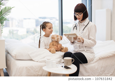 Pediatrician with tablet sitting next to girl on white bed 107631842