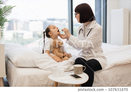 Young girl sitting on bed while doctor examining her throat Young girl sitting on bed while doctor examining her throat 107631870