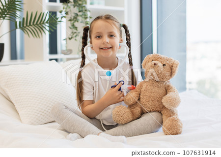 Smiling girl vaccinating teddy bear with toy syringe on bed Smiling girl vaccinating teddy bear with toy syringe on bed 107631914