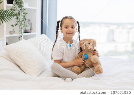 Pretty kid with toy stethoscope examining teddy bear in bed Pretty kid with toy stethoscope examining teddy bear in bed 107631920