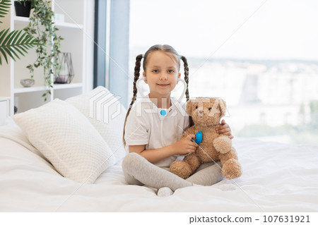 Pretty kid with toy stethoscope examining teddy bear in bed Pretty kid with toy stethoscope examining teddy bear in bed 107631921