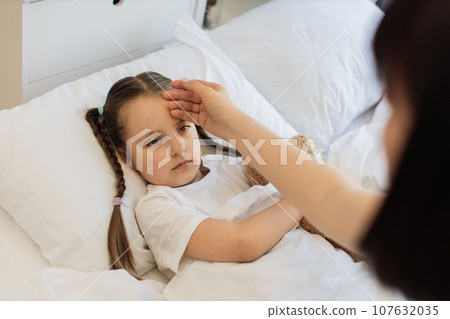 Sad ill girl lying in bed while mother checking temperature 107632035