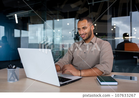 Young successful businessman working inside the office with a laptop, satisfied with the results of the achievement, the man is smiling, typing on the keyboard. Young successful businessman working inside the office with a laptop, satisfied with the results of the achievement, the man is smiling, typing on the keyboard. 107633267