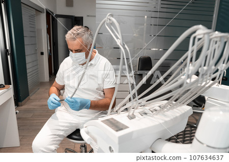 A dentist sits behind a dental chair and prepares tools for dental treatment. A senior, experienced male doctor works in a dental treatment and prosthetics center. A dentist sits behind a dental chair and prepares tools for dental treatment. A senior, experienced male doctor works in a dental treatment and prosthetics center. 107634637