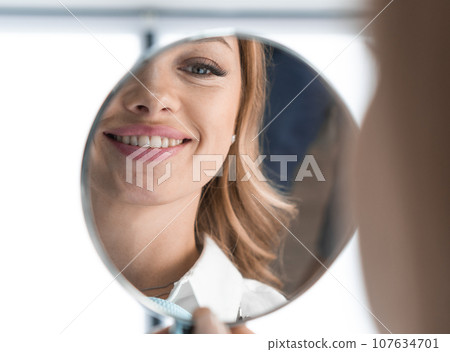 In mirror can see distinct improvements in condition teeth after visiting dentist. Woman is glad that can smile without complexes because her wonderful smile. After teeth whitening, implant placement 107634701