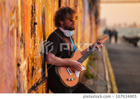 Hipster street musician in black playing electric guitar in the street on sunset leaning on a painted wall Hipster street musician in black playing electric guitar in the street on sunset leaning on a painted wall 107634805