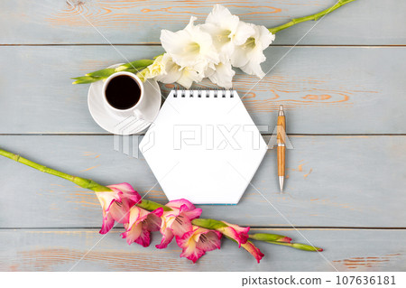 Composition with opened blank notepad and pen, cup of black coffee, white and pink gladiolus flower on blue wooden table. Festive office desktop concept. Morning coffee cup. 107636181