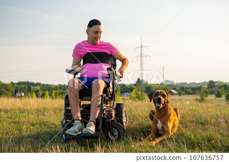 A happy disabled man with a wheelchair and his service dog at the park during sunset. A happy disabled man with a wheelchair and his service dog at the park during sunset. 107636757