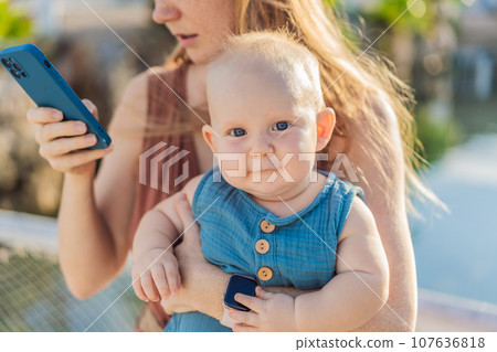 Mom holds her adorable baby while holding a smartphone 107636818
