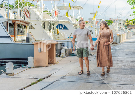 A happy, mature couple over 40, enjoying a leisurely walk on the waterfront, their joy evident as they embrace the journey of pregnancy later in life 107636861