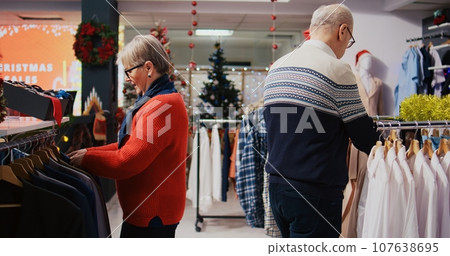 Octogenarian clients browsing through clothing racks in festive ornate fashion shop during winter holiday season. Senior couple happy after finding colorful blazers to gift at Xmas family gathering 107638695