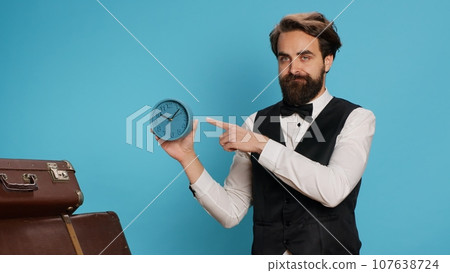 Doorman or bellhop, hotel staff checks time in studio while holding indoor clock to see the hour. Trying to be on time, young adult employee stands in formal attire with watch in hand. Doorman or bellhop, hotel staff checks time in studio while holding indoor clock to see the hour. Trying to be on time, young adult employee stands in formal attire with watch in hand. 107638724