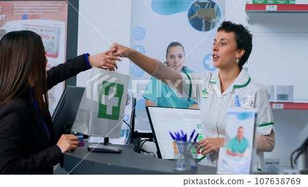 Shopper in chemist store at checkout counter using credit card to purchase pharmaceutical products. Friendly client doing contactless paying in pharmacy after finding needed ailment treatment pills 107638769