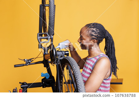 African American female cyclist carefully examines and repairs bike components with specialized tools and lubricant for smooth performance. Black woman diligently greasing and servicing bicycle chain. African American female cyclist carefully examines and repairs bike components with specialized tools and lubricant for smooth performance. Black woman diligently greasing and servicing bicycle chain. 107638788