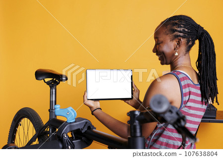 Energetic black woman standing near faulty bicycle and horizontally holding tablet with blank chromakey mockup template. Young female carrying digital device displaying whitescreen. Energetic black woman standing near faulty bicycle and horizontally holding tablet with blank chromakey mockup template. Young female carrying digital device displaying whitescreen. 107638804