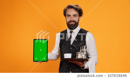 Friendly waiter shows tablet with greenscreen on camera, presenting blank copyspace mockup template. Experienced butler in suit holding device with isolated empty display and food tray. 107638846