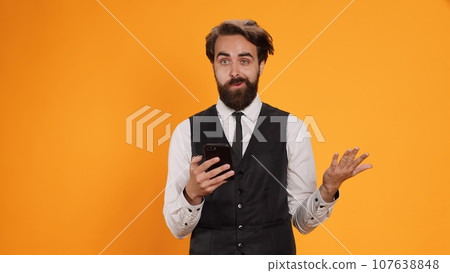 Restaurant employee messaging on duty and using phone at work, serving clients with meal orders. Classy waiter standing against yellow background in studio, checking smartphone social media apps. 107638848