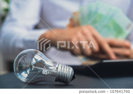 Electric light bulb. Man hands counting expenses bills on electricity banknotes of euro cash accounting on calculator. Close up of hands unrecognizable Businessman. High prices for energy. Save up Electric light bulb. Man hands counting expenses bills on electricity banknotes of euro cash accounting on calculator. Close up of hands unrecognizable Businessman. High prices for energy. Save up 107641700