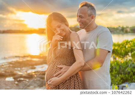 A happy, mature couple over 40, enjoying a leisurely walk on the waterfront On the Sunset, their joy evident as they embrace the journey of pregnancy later in life 107642262