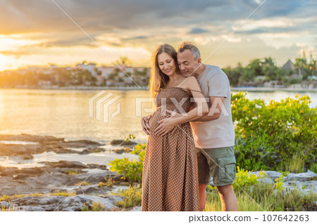 A happy, mature couple over 40, enjoying a leisurely walk on the waterfront On the Sunset, their joy evident as they embrace the journey of pregnancy later in life 107642263