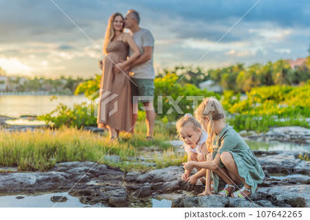A happy, mature couple over 40 with their two daughters enjoying a leisurely walk on the waterfront, their joy evident as they embrace the journey of pregnancy later in life 107642265