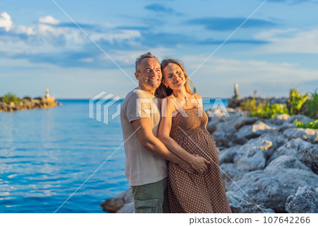 A happy, mature couple over 40, enjoying a leisurely walk on the waterfront On the Sunset, their joy evident as they embrace the journey of pregnancy later in life 107642266
