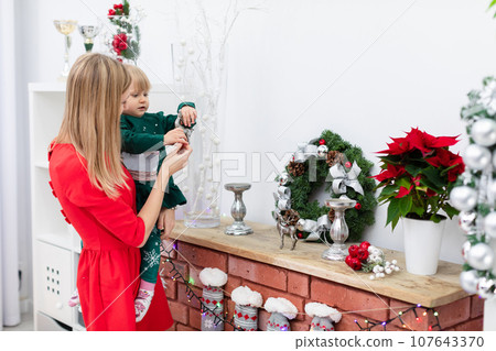 Mom holds her little daughter in her arms and shows her the Christmas decorations Mom holds her little daughter in her arms and shows her the Christmas decorations 107643370