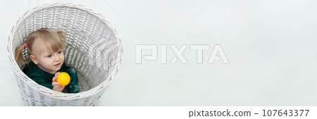 A small child plays with a yellow ball while sitting in a large wicker basket 107643377