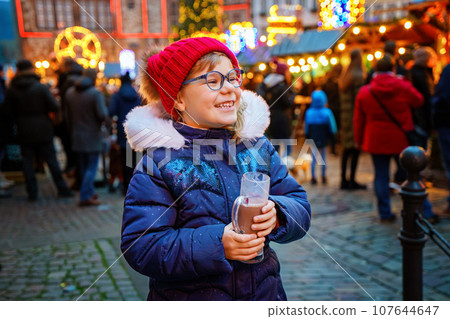 Little cute preschool girl drinking hot children punch or chocolate on German Christmas market. Happy child on traditional family market in Germany, Laughing kid in colorful winter clothes 107644647