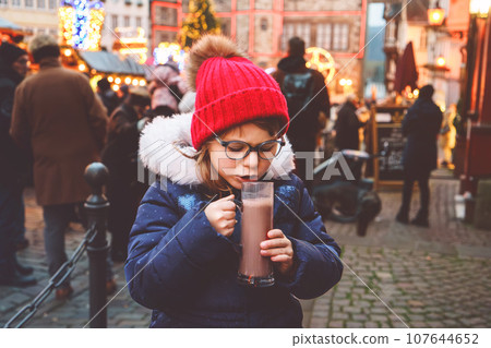 Little cute preschool girl drinking hot children punch or chocolate on German Christmas market. Happy child on traditional family market in Germany, Laughing kid in colorful winter clothes 107644652
