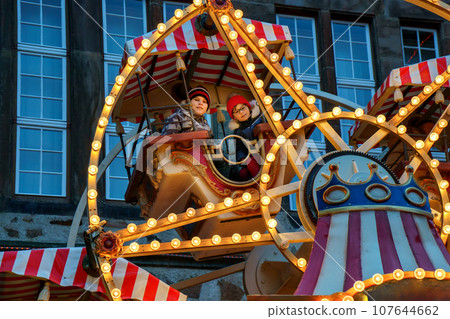 Little preschool girl and school boy riding on ferris wheel carousel horse at Christmas funfair or market, outdoors. Two happy children having fun on traditional family xmas market in Germany Little preschool girl and school boy riding on ferris wheel carousel horse at Christmas funfair or market, outdoors. Two happy children having fun on traditional family xmas market in Germany 107644662