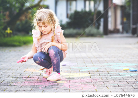 Little preschool girl painting with colorful chalks flowers on ground on backyard. Positive happy toddler child drawing and creating pictures on asphalt. Creative outdoors children activity in summer. 107644676