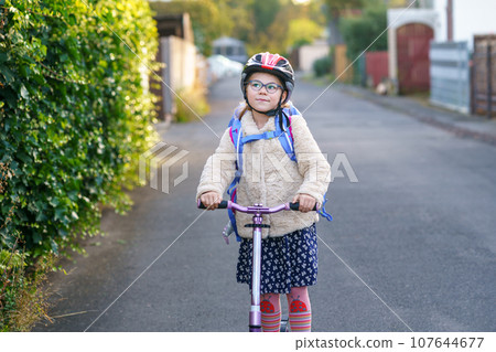 Little girl rides a kick scooter on her way back to school. Cute child with backpack and with safety helmet. Happy schoolchild. Little girl rides a kick scooter on her way back to school. Cute child with backpack and with safety helmet. Happy schoolchild. 107644677